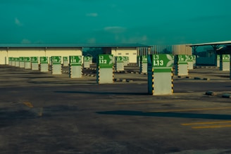 An empty parking lot with rows of parking spaces marked by white pillars featuring green signs. The signs display parking spot numbers and letters, with a stylized leaf emblem. Surrounding the area are covered structures that provide shade, and the scene is under a turquoise sky.
