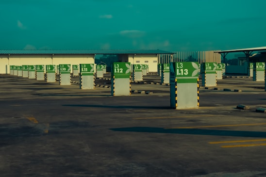 An empty parking lot with rows of parking spaces marked by white pillars featuring green signs. The signs display parking spot numbers and letters, with a stylized leaf emblem. Surrounding the area are covered structures that provide shade, and the scene is under a turquoise sky.