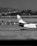 A black and white photograph of an airport scene featuring the tail of an airplane on the tarmac. The image captures a backdrop of hills and a cluster of buildings in the distance. Various airport vehicles are lined up near the aircraft.