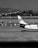 A black and white photograph of an airport scene featuring the tail of an airplane on the tarmac. The image captures a backdrop of hills and a cluster of buildings in the distance. Various airport vehicles are lined up near the aircraft.