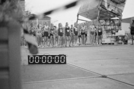 A group of runners is standing in a line, seemingly preparing for a race. The focus is on a digital timer on the ground displaying all zeros. The background appears to be an urban setting with concrete surfaces and a structure covered in graffiti.