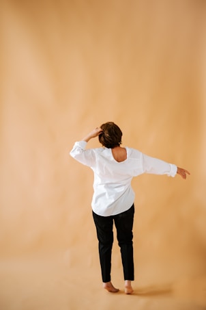 A person stands barefoot on a plain beige background. They are wearing a white long-sleeved shirt and black pants. Their right arm is extended outward while their left hand touches their head, creating a relaxed posture.