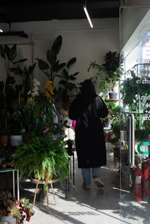 A person wearing a long black coat is standing inside a plant shop surrounded by a variety of potted plants. The space is well-lit with natural and artificial light, casting shadows and highlights on the plants and floor. There are large leafy plants on stands and shelves, creating a lush, green atmosphere. The glass door leading into the shop is partially open.