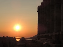 A stunning sunset over a body of water, partially obscured by an ancient structure with tall columns. The sun casts warm golden hues over the landscape, creating a dramatic silhouette of the ruins and the distant hills.