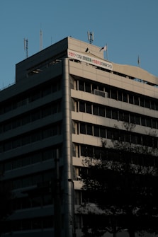 The image displays a multi-story office building with a modern architectural design. The building has a steel structure with horizontal rows of windows and a column-like structure on one corner. There are antennas and a flagpole on the rooftop. The lighting suggests it may be late afternoon or early morning.