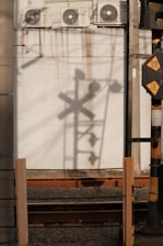 The image features a shadow of a railway crossing signal cast on a white, stained wall with multiple air conditioning units at the top. Below the signal, railway tracks run horizontally. The industrial setting is complemented by a vertical post with caution signs.
