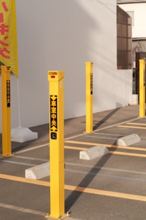 Several yellow parking bollards with black and red text stand in a neat arrangement next to white parking blocks. A yellow sign with red text is partially visible on the left, mounted on a pole. Sunlight casts shadows across the gray pavement and the white building wall in the background.