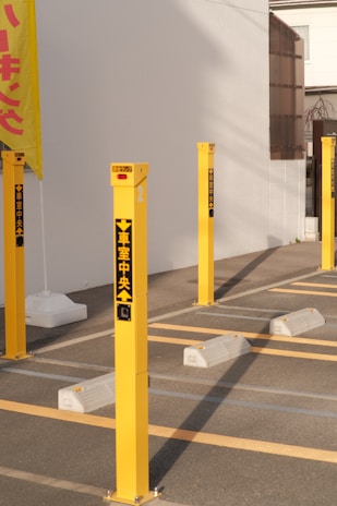 Several yellow parking bollards with black and red text stand in a neat arrangement next to white parking blocks. A yellow sign with red text is partially visible on the left, mounted on a pole. Sunlight casts shadows across the gray pavement and the white building wall in the background.