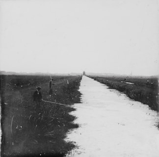 A black and white image of a rural landscape with a canal running through the center. Two people are visible, one standing on either side of the canal, both appearing to be fishing. In the background, a windmill is visible on the horizon, surrounded by flat fields.
