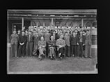 A group of young men and teachers are posing for a formal group photograph in front of a school building. The individuals are dressed in suits and ties, suggesting it is a school or academic setting.