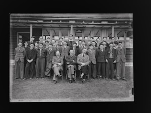 A group of young men and teachers are posing for a formal group photograph in front of a school building. The individuals are dressed in suits and ties, suggesting it is a school or academic setting.