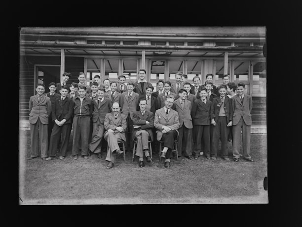 A group of young men and teachers are posing for a formal group photograph in front of a school building. The individuals are dressed in suits and ties, suggesting it is a school or academic setting.