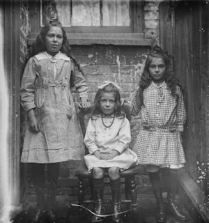 Three young girls pose in a small alleyway between brick walls. Two stand while one sits in a wooden chair. They wear dresses with structured collars and long socks with boots, reflecting early 20th-century fashion. The scene appears to be in an urban setting, with a window visible in the background.