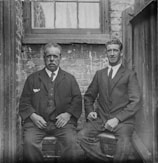 Two men in formal suits are seated side by side on stools against an old brick wall. The man on the left has a serious expression and wears a dark suit with a white pocket square. The man on the right appears more relaxed and is also dressed in a dark suit and tie. They are positioned in front of a window with lace curtains inside an alleyway-like setting.