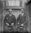 Two men in formal suits are seated side by side on stools against an old brick wall. The man on the left has a serious expression and wears a dark suit with a white pocket square. The man on the right appears more relaxed and is also dressed in a dark suit and tie. They are positioned in front of a window with lace curtains inside an alleyway-like setting.