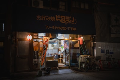 A small cozy storefront with a dark blue awning adorned with Japanese characters. Warm lighting spills out onto the street, creating a welcoming atmosphere. Two bicycles are parked in front, and traditional red lanterns hang on either side of the entrance. There are colorful banners with text and graphics near the doorway.