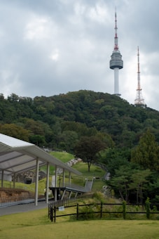 A tall communication tower rises prominently from a wooded hill, surrounded by dense, lush greenery. The foreground features a modern, open-air structure with a sleek white roof, set amidst a well-maintained grassy area with a wooden fence. The sky is overcast with thick, gray clouds.