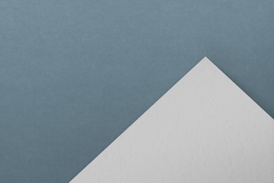 A minimalist composition with a white triangular piece of paper placed against a textured, muted blue background. The arrangement creates a contrast between the smooth, clean edges of the paper and the soft, even surface of the background.