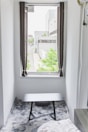 A small room features a window with open curtains, offering a view of a modern building complex with greenery outside. Inside, a black table sits on a gray, textured rug, and a part of a bed is visible in the corner.