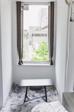 A small room features a window with open curtains, offering a view of a modern building complex with greenery outside. Inside, a black table sits on a gray, textured rug, and a part of a bed is visible in the corner.