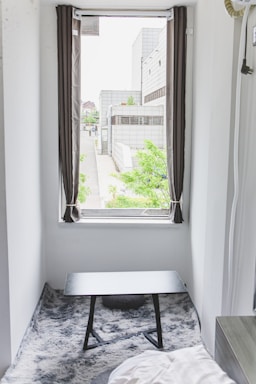 A small room features a window with open curtains, offering a view of a modern building complex with greenery outside. Inside, a black table sits on a gray, textured rug, and a part of a bed is visible in the corner.