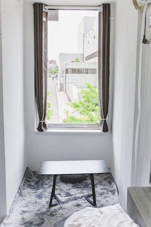 A small room features a window with open curtains, offering a view of a modern building complex with greenery outside. Inside, a black table sits on a gray, textured rug, and a part of a bed is visible in the corner.