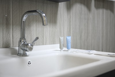 A clean and modern bathroom sink with a shiny metal faucet. Two small tubes of toiletries, one gray and one light blue, are placed on the counter next to the faucet. The background features a textured, patterned wall with neutral tones.