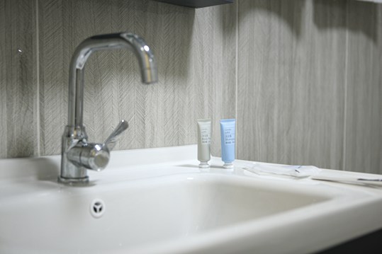 A clean and modern bathroom sink with a shiny metal faucet. Two small tubes of toiletries, one gray and one light blue, are placed on the counter next to the faucet. The background features a textured, patterned wall with neutral tones.