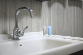 A clean and modern bathroom sink with a shiny metal faucet. Two small tubes of toiletries, one gray and one light blue, are placed on the counter next to the faucet. The background features a textured, patterned wall with neutral tones.