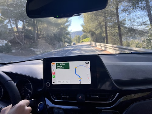 A car interior with a navigation system screen displaying a route. The driver's hand is on the steering wheel. Through the windshield, a road bordered by trees and a metal guardrail is visible. The day is sunny with clear skies.