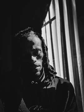 A person with dreadlocks is sitting in a shadowed room next to a window with vertical blinds. The light casts dramatic shadows across their face, creating a high contrast black and white effect.