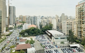 An urban cityscape with high-rise buildings and a densely built environment. Various architectural styles are evident, with a mix of modern apartment blocks and older structures. A busy street lined with numerous parked cars can be seen, with trees and vegetation providing some green space amid the concrete.