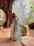 A serene woman wearing a flowing ceramic-inspired dress, standing in a sunlit room filled with plants.
