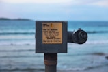A pay-to-use telescope is positioned on a beach with a view of the sea. It features a sign with instructions and information about usage, mounted on a rusty metal stand. The ocean in the background is calm with gentle waves under a partly cloudy sky.