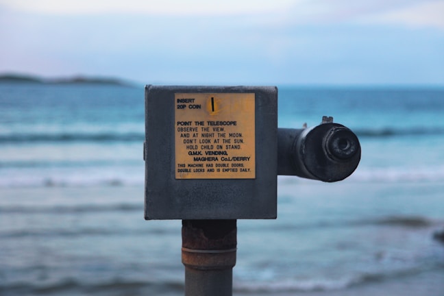 A pay-to-use telescope is positioned on a beach with a view of the sea. It features a sign with instructions and information about usage, mounted on a rusty metal stand. The ocean in the background is calm with gentle waves under a partly cloudy sky.