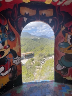 A colorful, painted archway frames a scenic landscape of mountains and lush greenery. The artwork on the archway features vibrant cartoonish characters with exaggerated expressions. Beyond the arch, the sky is partly cloudy with distant hills and a clear view of nature.