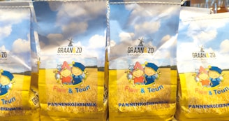 Packages of pancake mix featuring illustrations of two cartoon children in traditional attire against a backdrop of a wheat field and a blue sky with fluffy clouds.