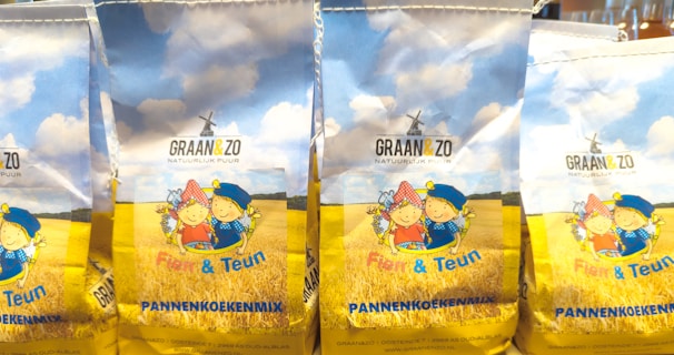 Packages of pancake mix featuring illustrations of two cartoon children in traditional attire against a backdrop of a wheat field and a blue sky with fluffy clouds.