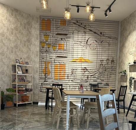 A cozy indoor space featuring a large wall mural with street and building illustrations in brown and yellow tones. There are multiple wooden tables and chairs in the room, arranged neatly. The space includes decorative lighting hanging from the ceiling, a bookshelf with various items including books and plants, and green plants placed in pots. The overall decor is modern and inviting, with touches of creativity and warmth.