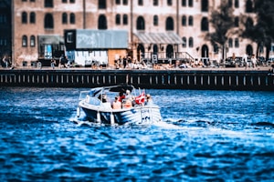 A small motorboat with several passengers, including two adults and a child, cruises on a river or canal. A Danish flag is visibly mounted at the back of the boat. The background features an industrial-style building with numerous arched windows, and people are gathered along the waterfront, some appearing to relax or socialize.
