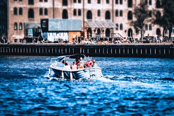 A small motorboat with several passengers, including two adults and a child, cruises on a river or canal. A Danish flag is visibly mounted at the back of the boat. The background features an industrial-style building with numerous arched windows, and people are gathered along the waterfront, some appearing to relax or socialize.