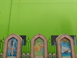 Three arched stone frames positioned in front of a bright green wall. Each frame contains an artistic illustration: the left one features traditional coffee pots, the middle showcases palm trees with dates, and the right displays a sailing ship with a map background. Metal fencing and windows are visible behind the frames.