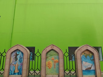 Three arched stone frames positioned in front of a bright green wall. Each frame contains an artistic illustration: the left one features traditional coffee pots, the middle showcases palm trees with dates, and the right displays a sailing ship with a map background. Metal fencing and windows are visible behind the frames.