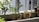 A small balcony garden with a planter containing leafy plants. Several smaller pots with succulents are lined up along the edge. The scene is well-lit with natural sunlight, casting soft shadows and highlighting the green foliage.