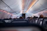 An airplane cabin interior with rows of seats filled with passengers. The view is from the back, looking towards the front where overhead luggage compartments are visible. Ambient lighting with an orange hue is present, illuminating the ceiling.