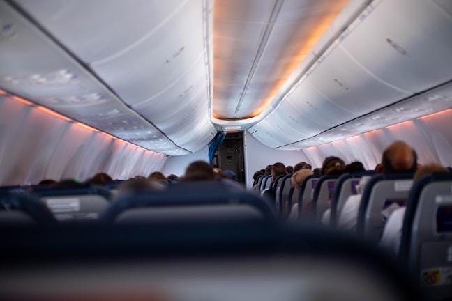 An airplane cabin interior with rows of seats filled with passengers. The view is from the back, looking towards the front where overhead luggage compartments are visible. Ambient lighting with an orange hue is present, illuminating the ceiling.