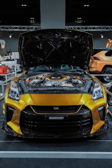 A bright yellow sports car with its hood open, showcasing a complex engine. The car is prominently displayed in an indoor setting with additional cars and a person visible in the background. The vehicle has a sleek design with detailed headlights and a large front grille.