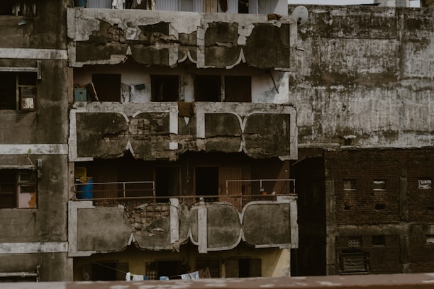 The image depicts an old, dilapidated apartment building with visible signs of wear and decay. The facade is cracked and peeling, with exposed concrete and paint chipping off. Windows are either missing or have improper coverings, and the balconies are poorly maintained with rusted railings. Various items, such as a blue barrel, are scattered on the balconies, adding to the sense of neglect.