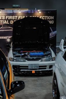 A modified white car is on display at an indoor automotive show, with the hood open to reveal a blue engine compartment. The setup includes a backdrop with partially visible signage and lighting fixtures. Nearby vehicles are partially visible, with one orange car in the foreground. The license plate features Japanese characters.