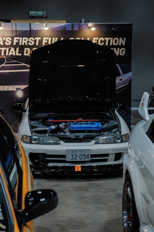 A modified white car is on display at an indoor automotive show, with the hood open to reveal a blue engine compartment. The setup includes a backdrop with partially visible signage and lighting fixtures. Nearby vehicles are partially visible, with one orange car in the foreground. The license plate features Japanese characters.